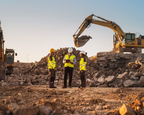 construction site with excavators on sunny day: diverse team of male and female specialists discussing real estate project. civil engineer, architect and investor talking about apartment building.