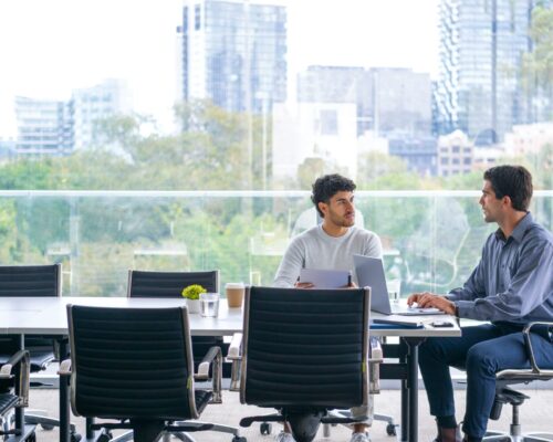 two businessmen working on a digital tablet and laptop computer in the office a board room or meeting room. there is a large window behind them